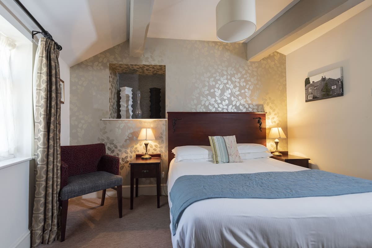 A hotel room with a bed, a chair, two bedside tables with lamps, a window with curtains, and a wall with a floral pattern and a picture.