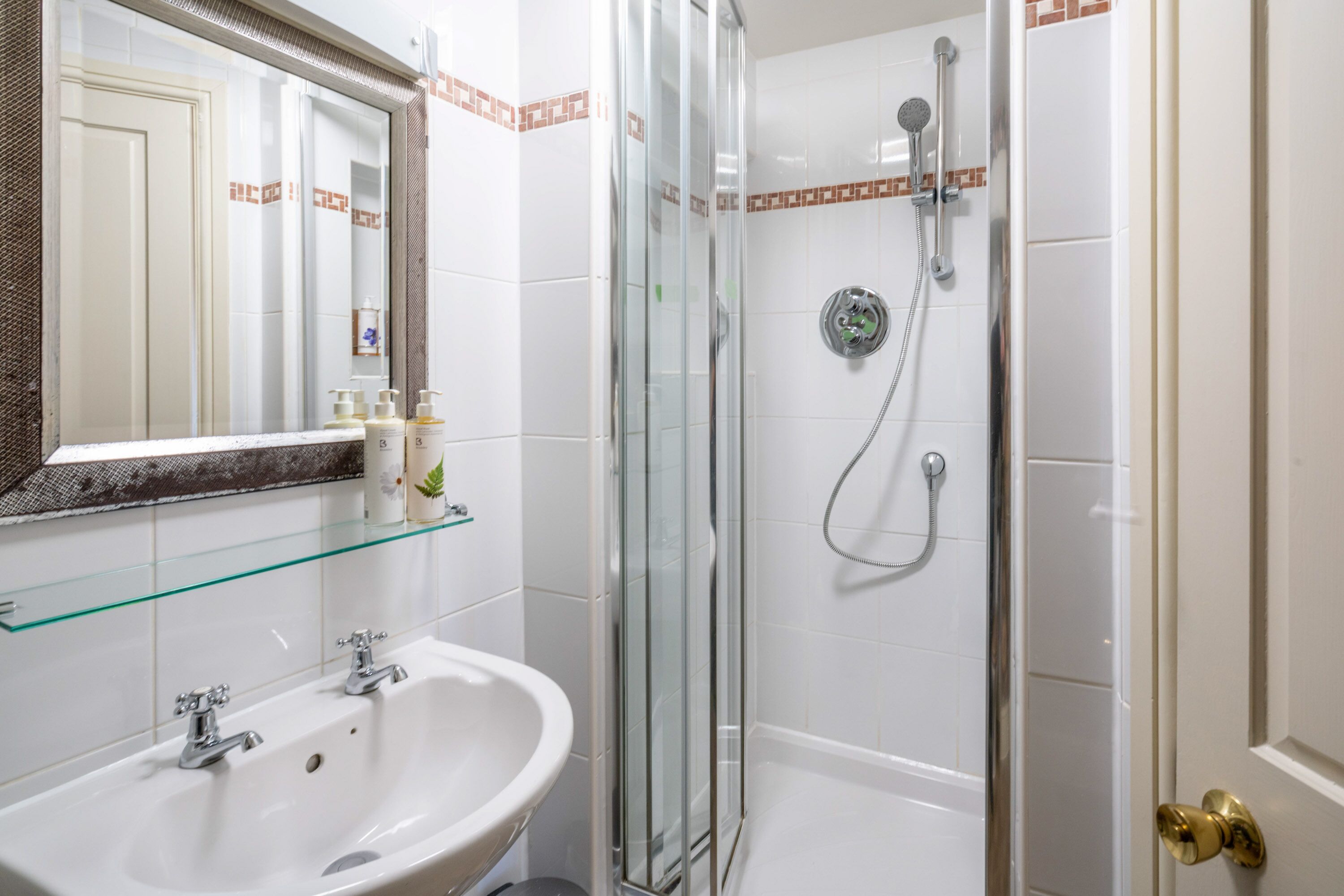 Un baño con cabina de ducha de vidrio, un lavabo blanco con dos grifos y un estante de vidrio sobre el lavabo.