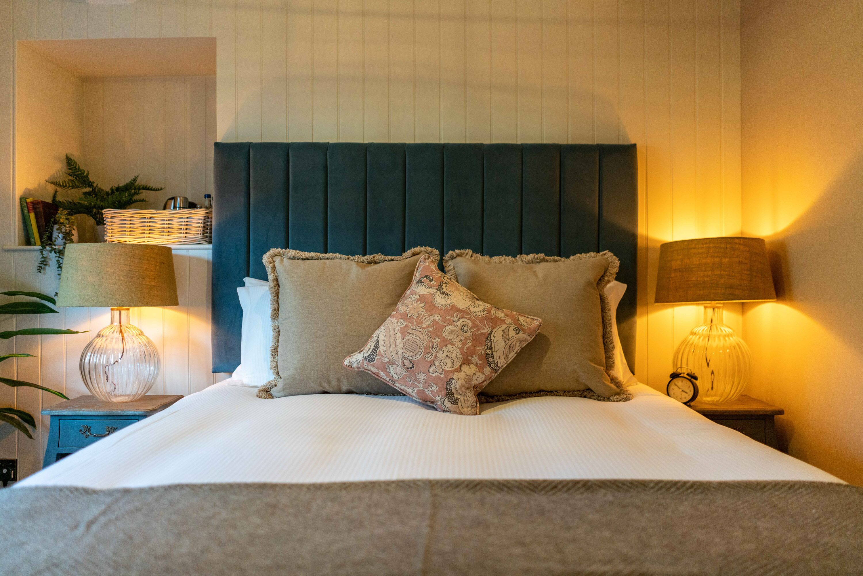 A neatly made bed with a dark headboard, two bedside lamps, and a small bedside table with a lamp and a clock.
