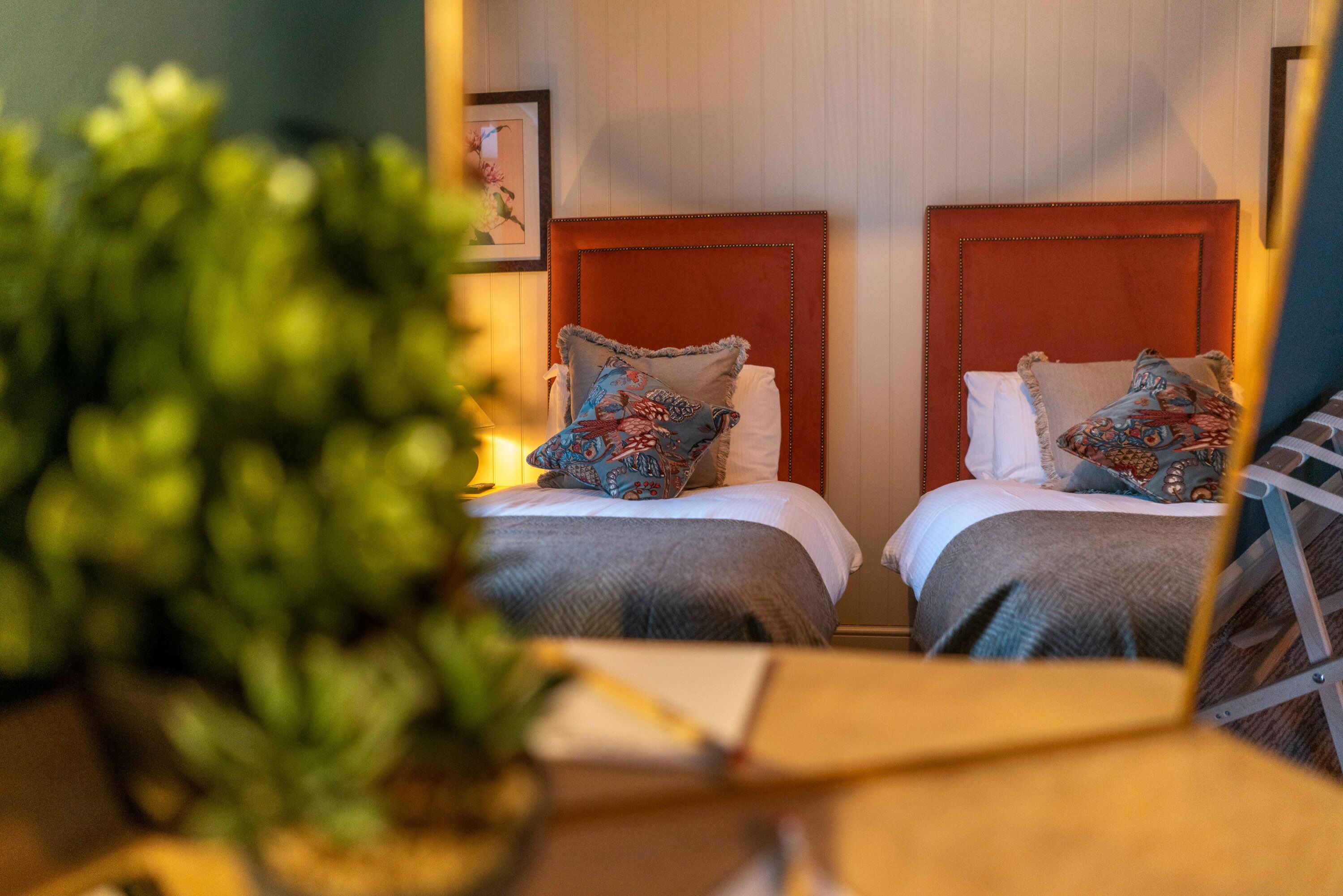 A hotel room with two beds, each with a red headboard, white bedding, and patterned pillows. There is a potted plant in the foreground.