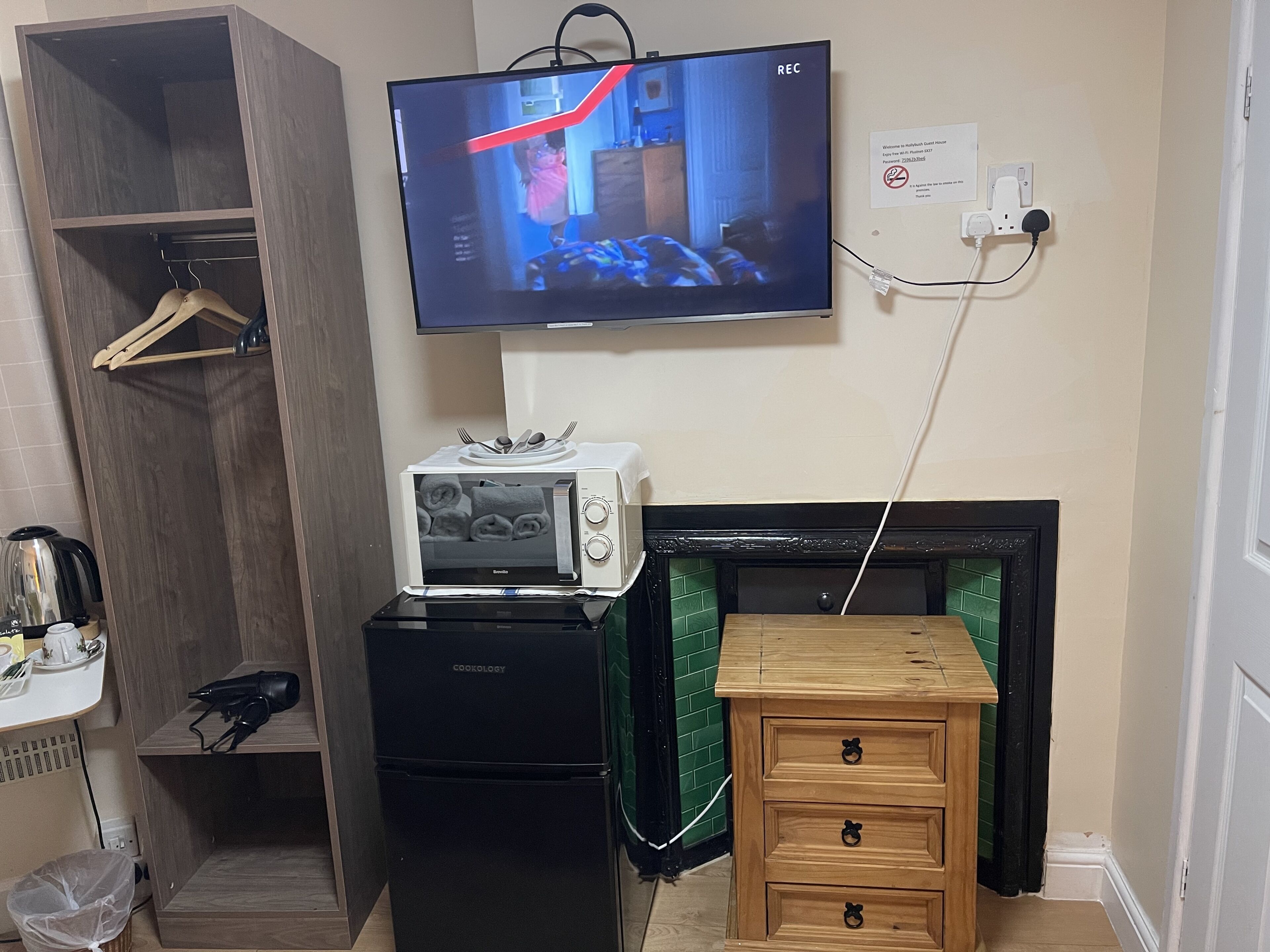 A compact room with a television mounted on the wall, a microwave, a small refrigerator, a wooden cabinet, and a fireplace.