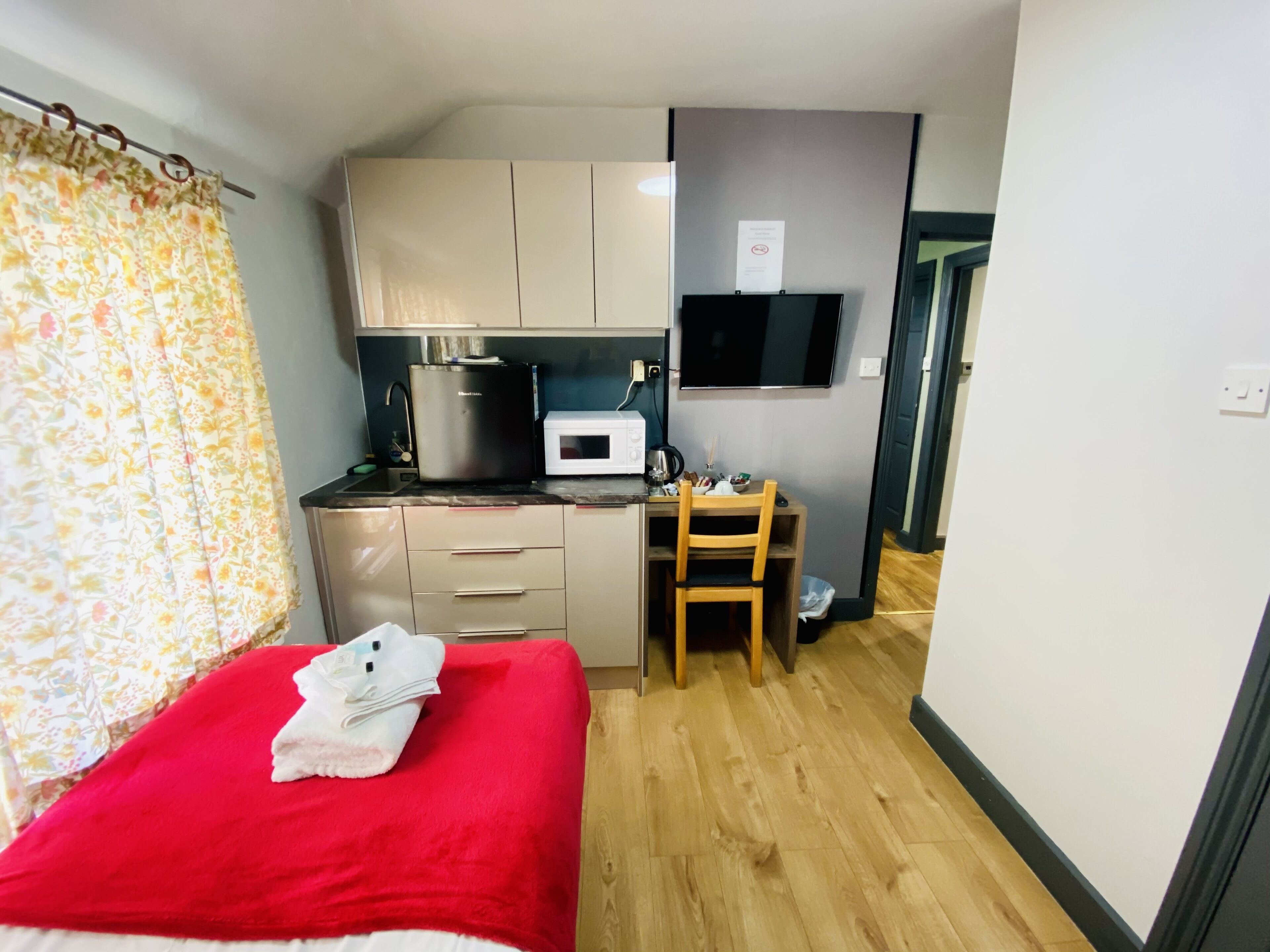 A compact room with a bed, a small kitchenette, a wooden dining table, and a TV mounted on the wall.