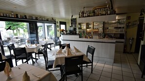 Un interior de restaurante con mesas preparadas para comer, una zona de bar con estantes llenos de botellas y vistas al exterior a través de grandes ventanales.
