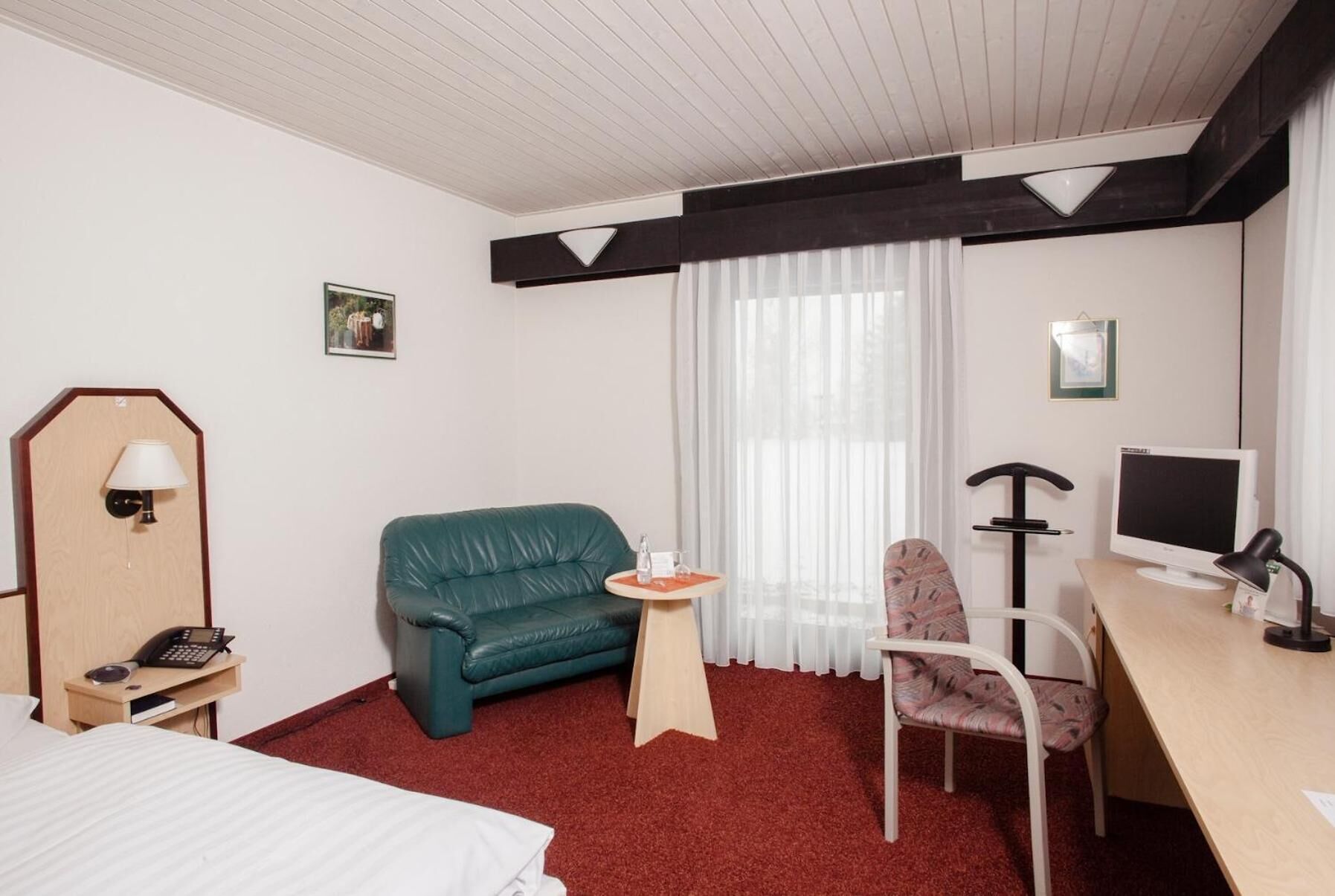 Habitació d'hotel amb un llit, un sofà verd, una taula petita amb un got, un escriptori amb un ordinador i una cadira.