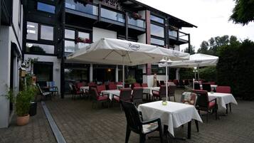 An outdoor dining area with tables and chairs, covered by white umbrellas, and a building with multiple floors and balconies.