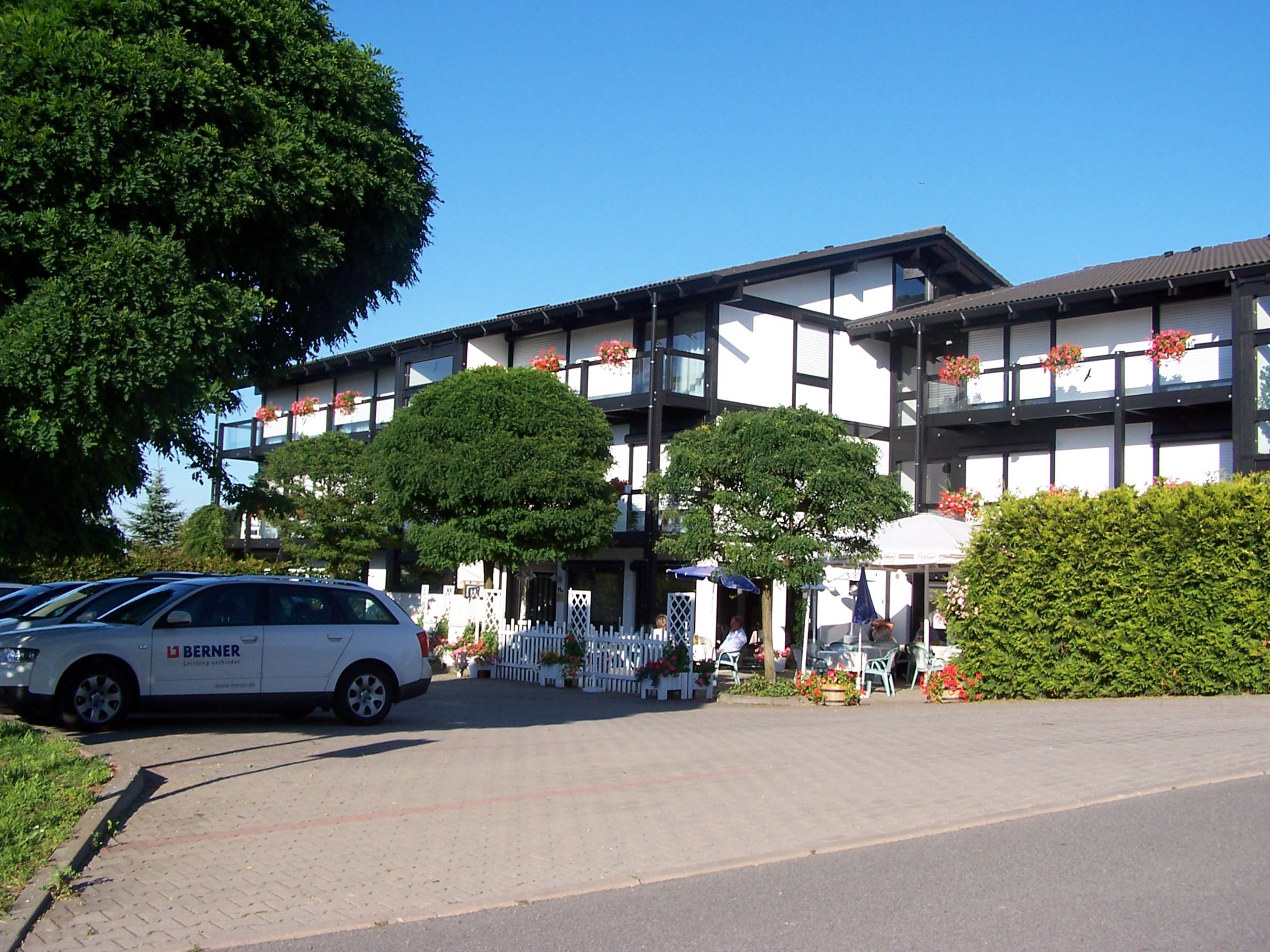 Un edificio con facciata bianca, numerose finestre e balconi adornati da fiori rossi. Davanti, alberi e una zona pavimentata con sedie e tavoli.