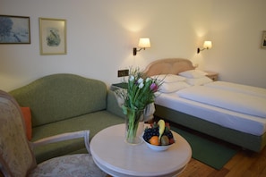 A room with a bed, a green sofa, a round table with a bowl of fruit and a vase of flowers, and wall-mounted lamps.