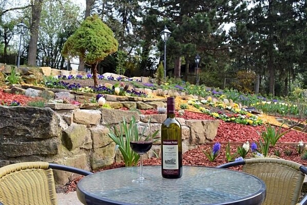 Una mesa de vidrio con una botella de vino y dos copas, rodeada de sillas de mimbre, en un jardín con diversas plantas y un muro de piedra.