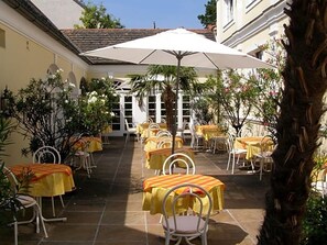 Un patio al aire libre con mesas y sillas, rodeado de plantas y un paraguas blanco.