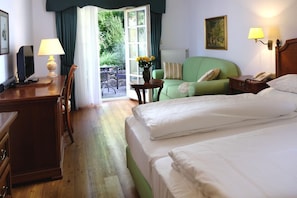 Ein Hotelzimmer mit Bett, Schreibtisch mit Lampe, Stuhl, kleinem Tisch mit Blumen und Blick ins Freie.