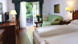 Habitación de hotel con cama, escritorio con lámpara, silla, pequeña mesa con flores y vistas al exterior.