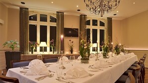 A long dining table set for a formal event, with chairs on both sides, large windows with curtains, and a chandelier. - Post, ZUR (Bonn)