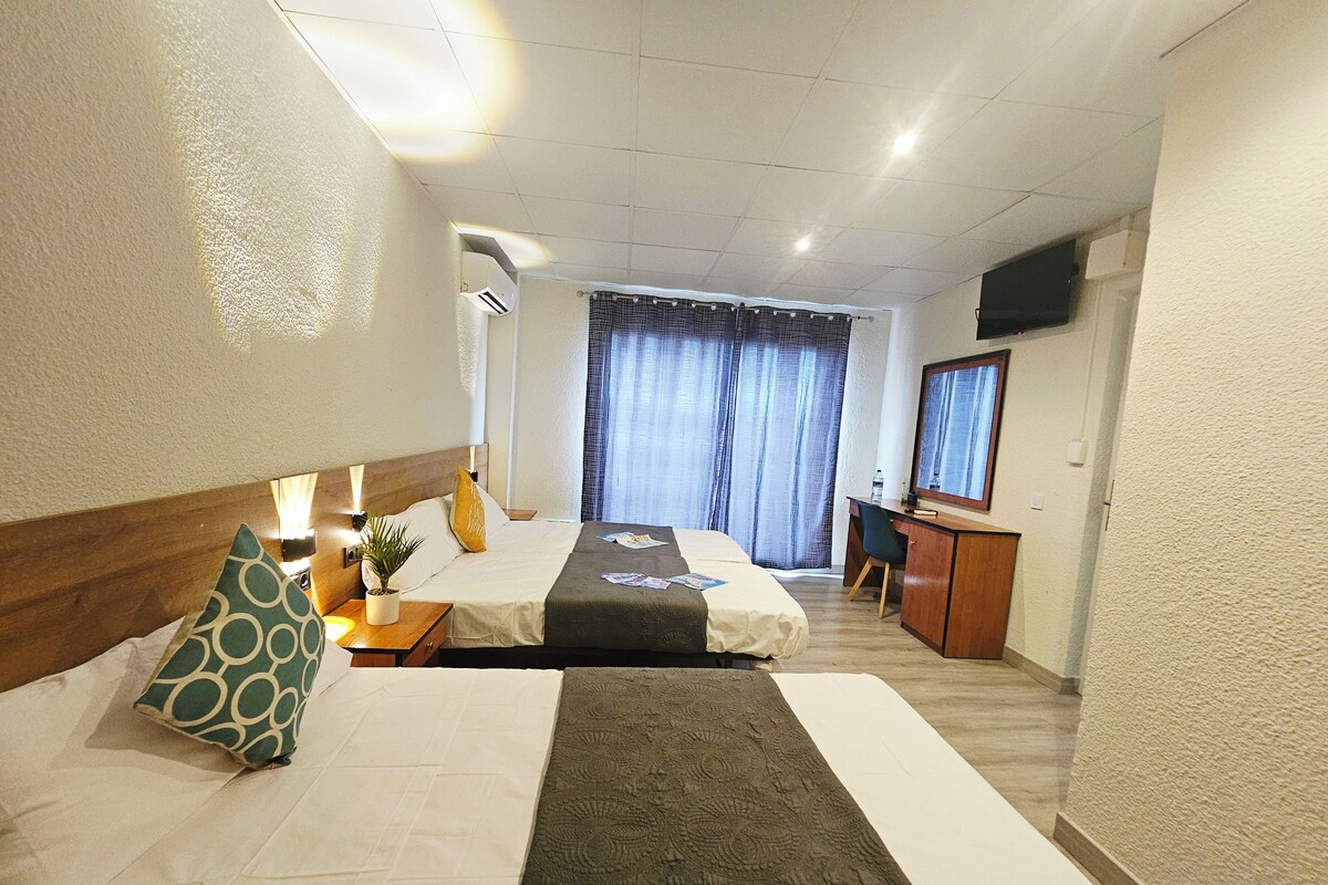Habitación de hotel con dos camas, un escritorio, una silla, televisión y un gran ventanal con cortinas.