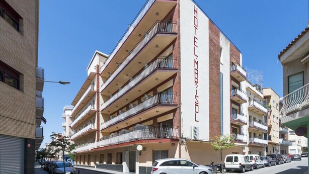 Un hotel de varios pisos con balcones en cada nivel, cielo despejado y coches aparcados en la calle.
