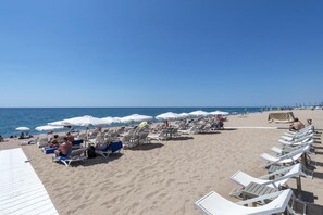 A beach with rows of white lounge chairs and umbrellas, a sandy beach, and the ocean in the background. - Marisol (Calella)