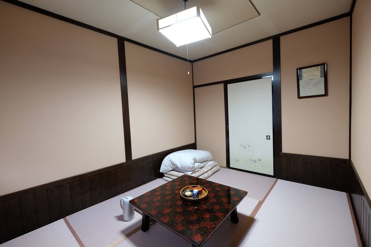 Une pièce avec un sol recouvert de tatamis, une petite table basse avec un plateau, un coussin et un avis encadré accroché au mur.