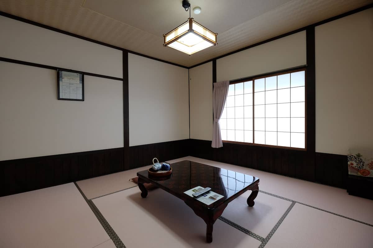 Une pièce de style japonais traditionnel, avec un sol recouvert de tatamis, une petite table en bois et une grande fenêtre agrémentée d’un rideau.