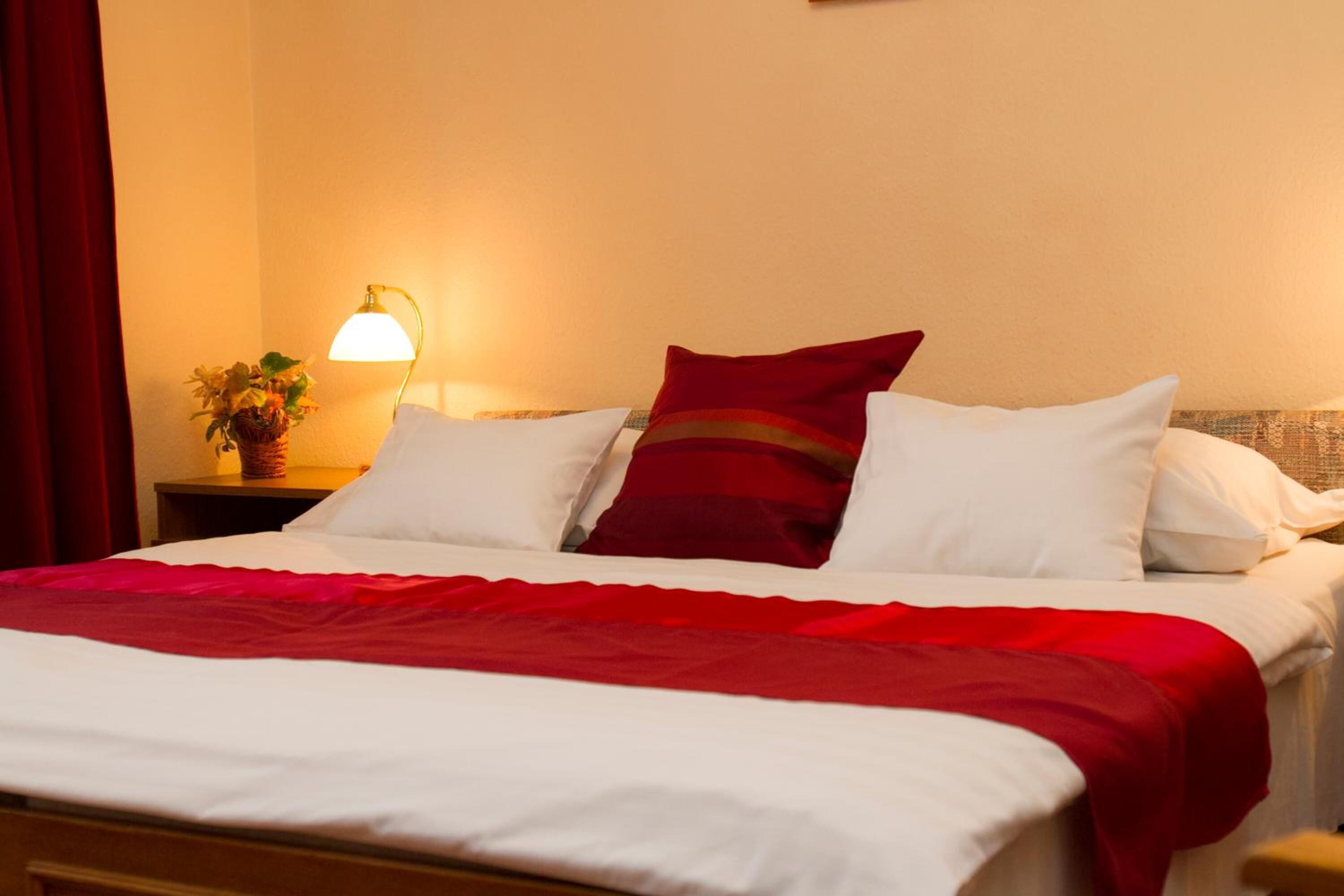 A neatly made bed with white and red bedding, a bedside lamp, and a small plant on a nightstand.