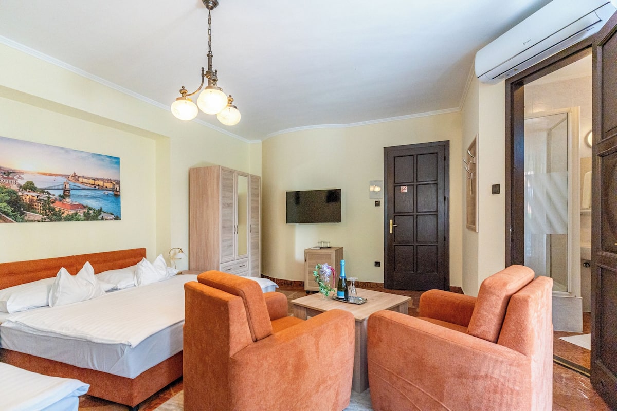 A hotel room with a bed, two orange armchairs, a small table with a bottle and glasses, a TV on the wall, and a painting of a bridge.