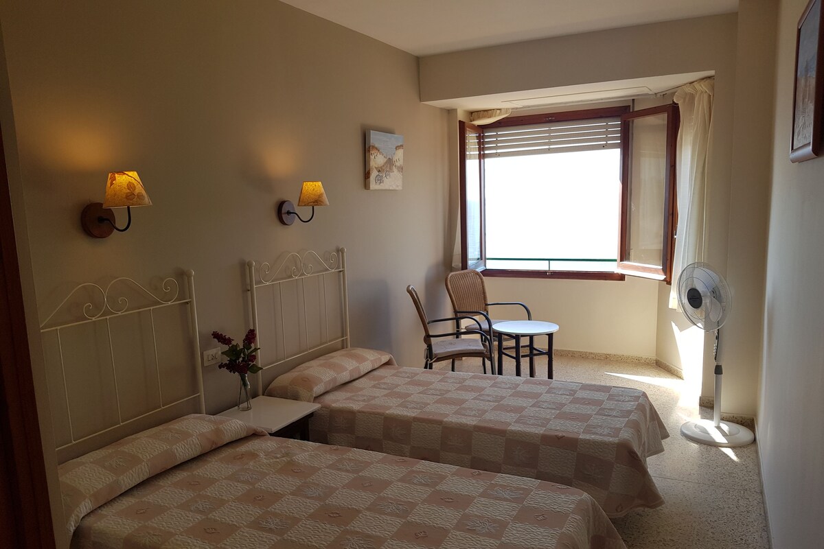 A room with two beds, a chair, a table, and a window with blinds.