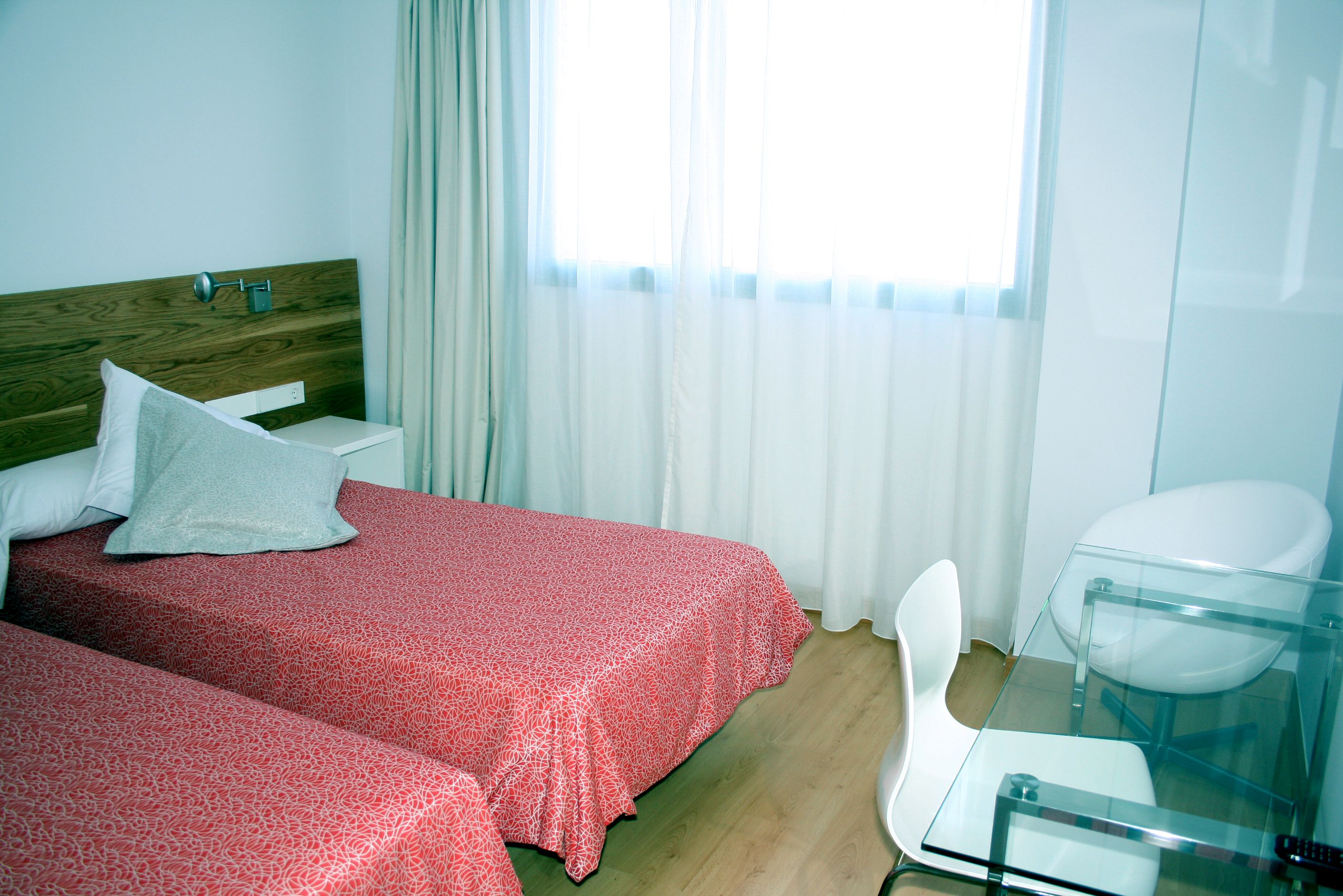 Habitación de hotel con dos camas, una mesa de vidrio, una silla blanca y una ventana con cortinas.