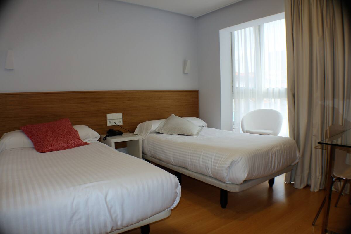 Habitación de hotel con dos camas, cabecera de madera, mesita de noche, una silla y ventana con cortinas.