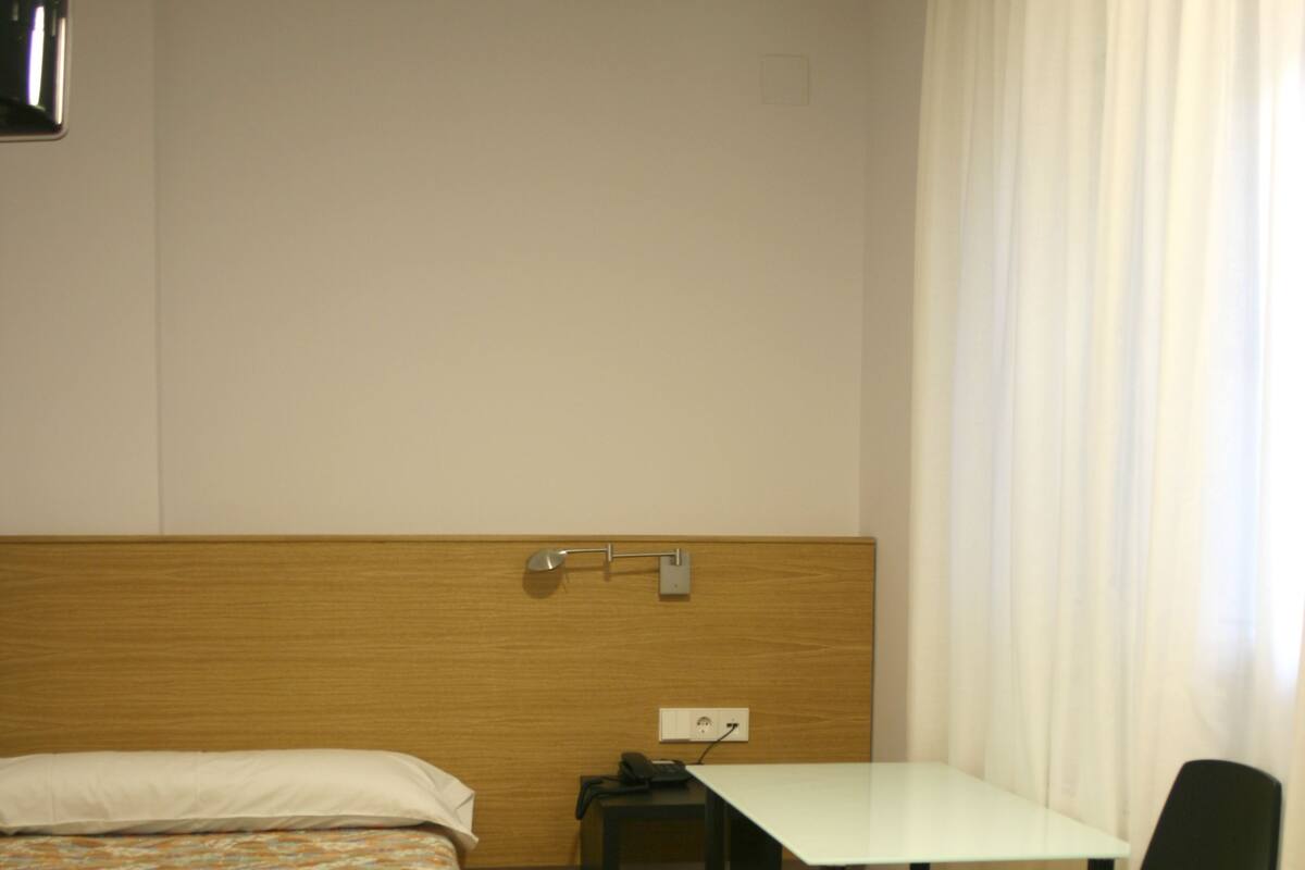 Una habitación de hotel con cama, escritorio, silla y ventana con cortinas.