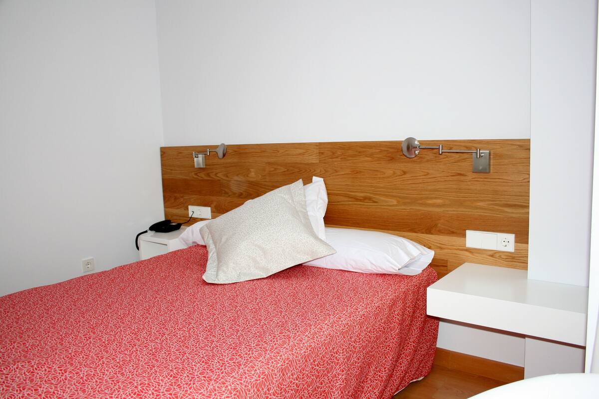 Una cama bien hecha con una colcha roja estampada, una almohada blanca y un cabecero de madera.