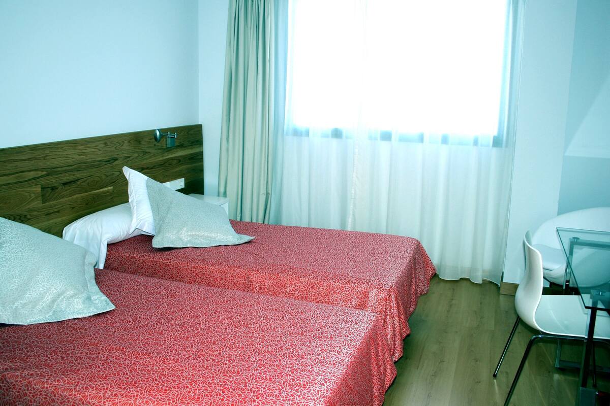 Habitación de hotel con cabecera de madera, dos camas con ropa de cama roja estampada, almohadas blancas, una mesita de cristal y una silla.