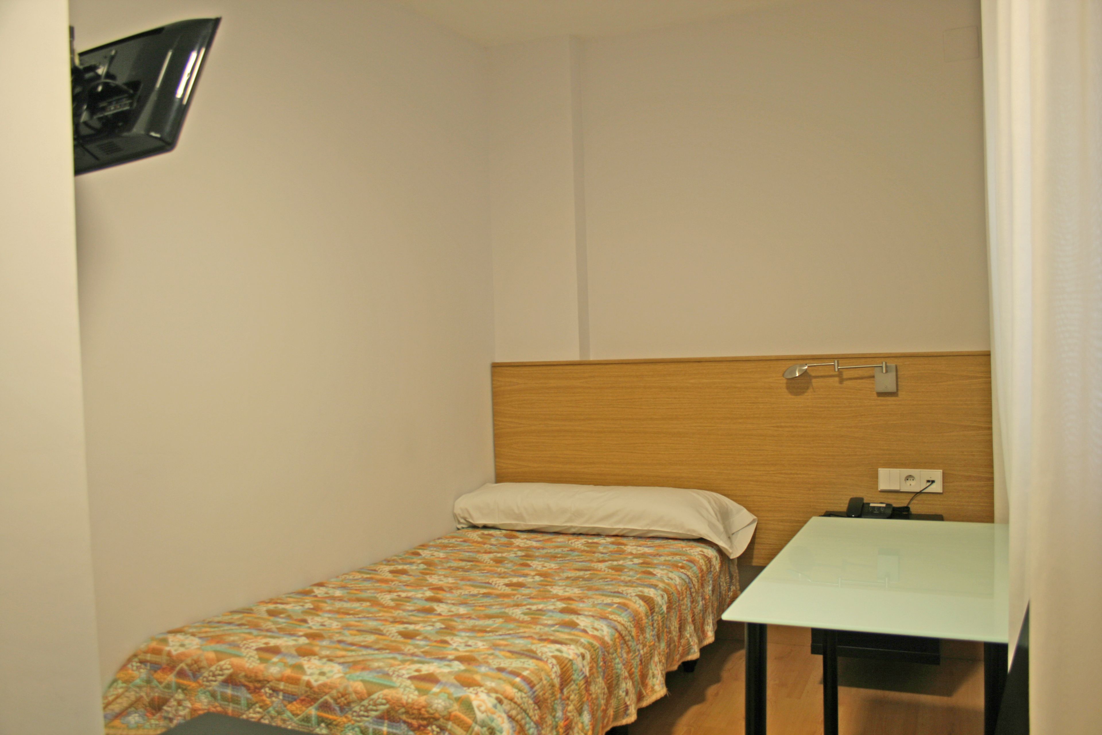 A small, single-bed room with a wooden headboard, a bedside table, and a power outlet on the wall.