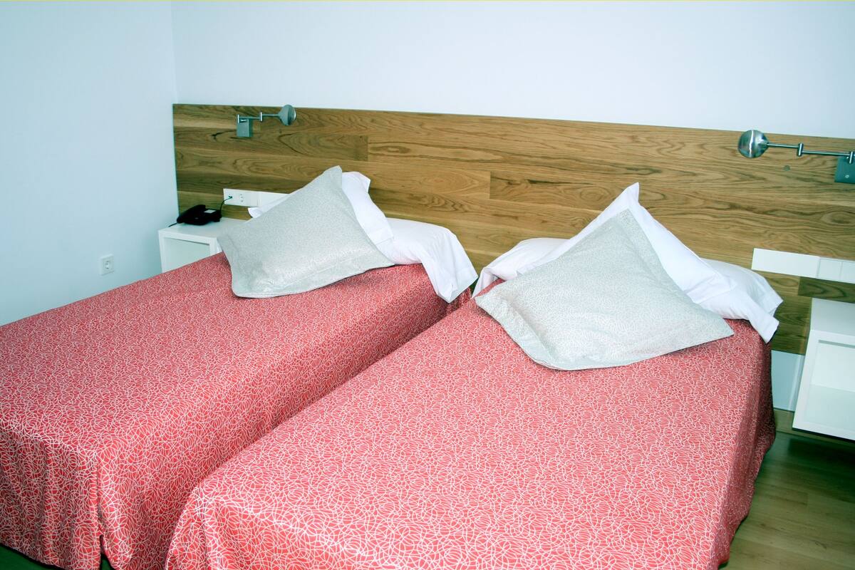 Cama doble con sábanas rojas estampadas y almohadas blancas, cabecera de madera y mesita de noche con una lámpara.