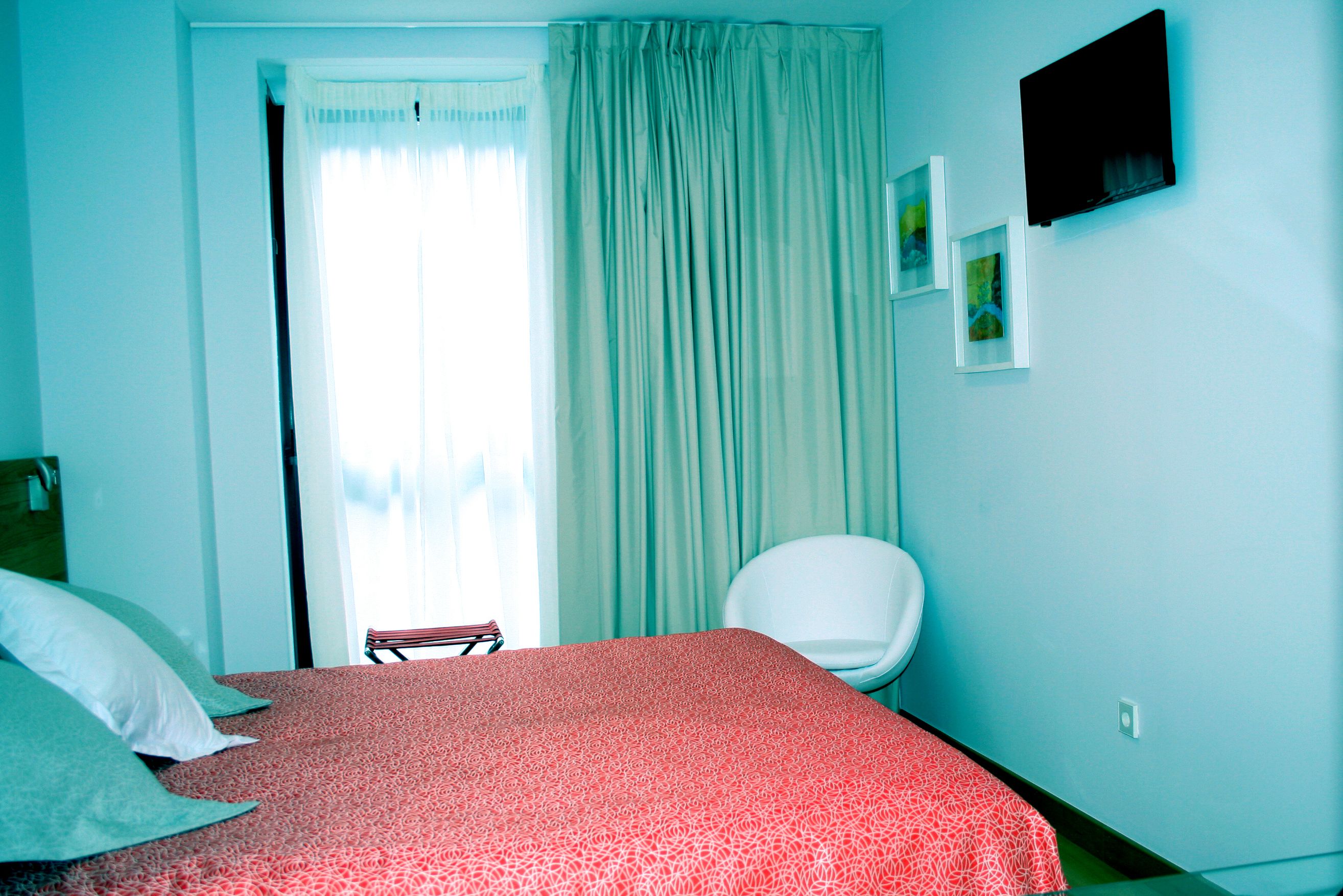 A bedroom with a bed, a chair, a window with curtains, and a wall-mounted TV.