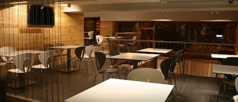 A modern restaurant with multiple tables and chairs, wooden accents, and a large window providing a view of the city.