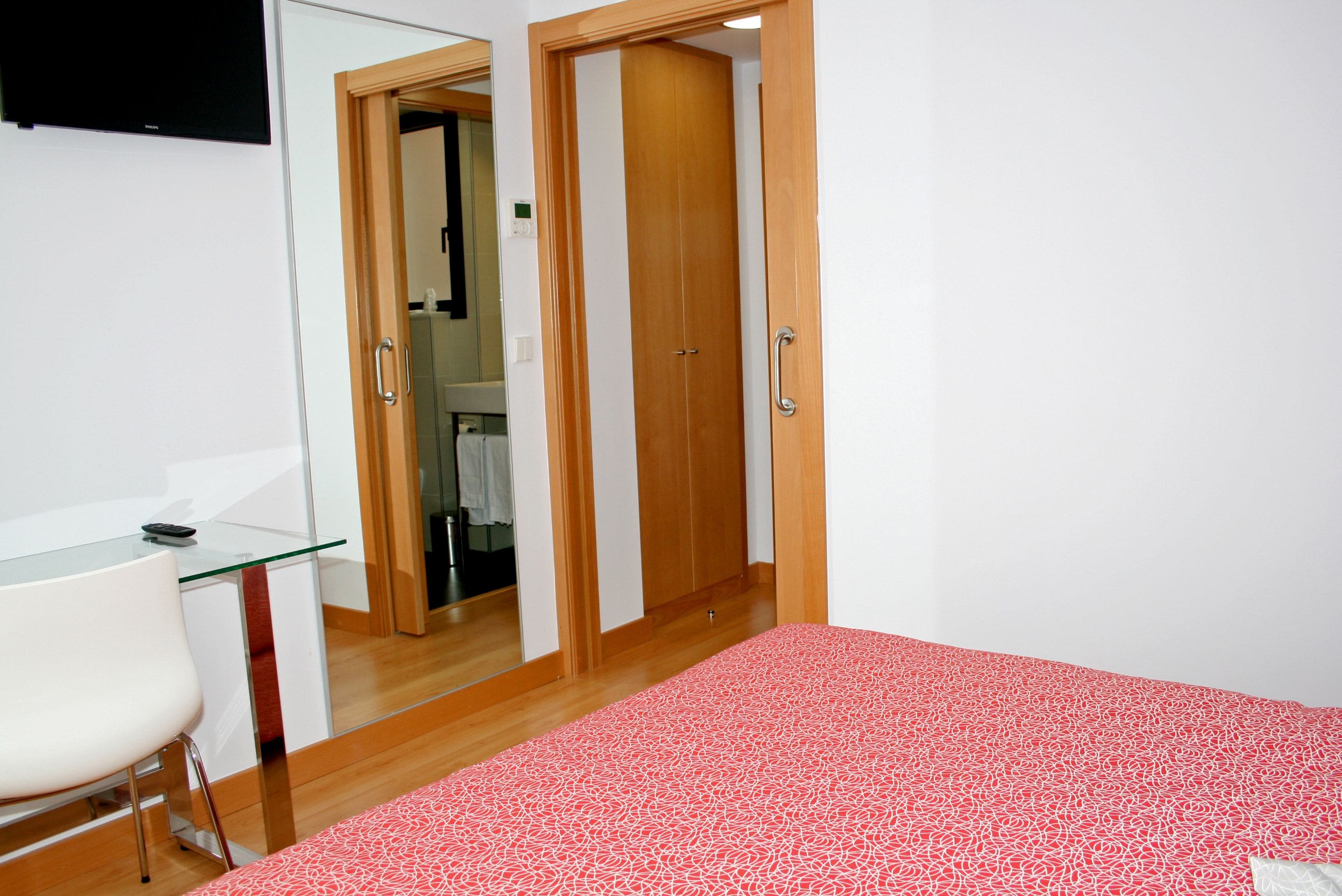 Un dormitorio con cama, escritorio y silla. Hay un armario de madera y un espejo en la pared.