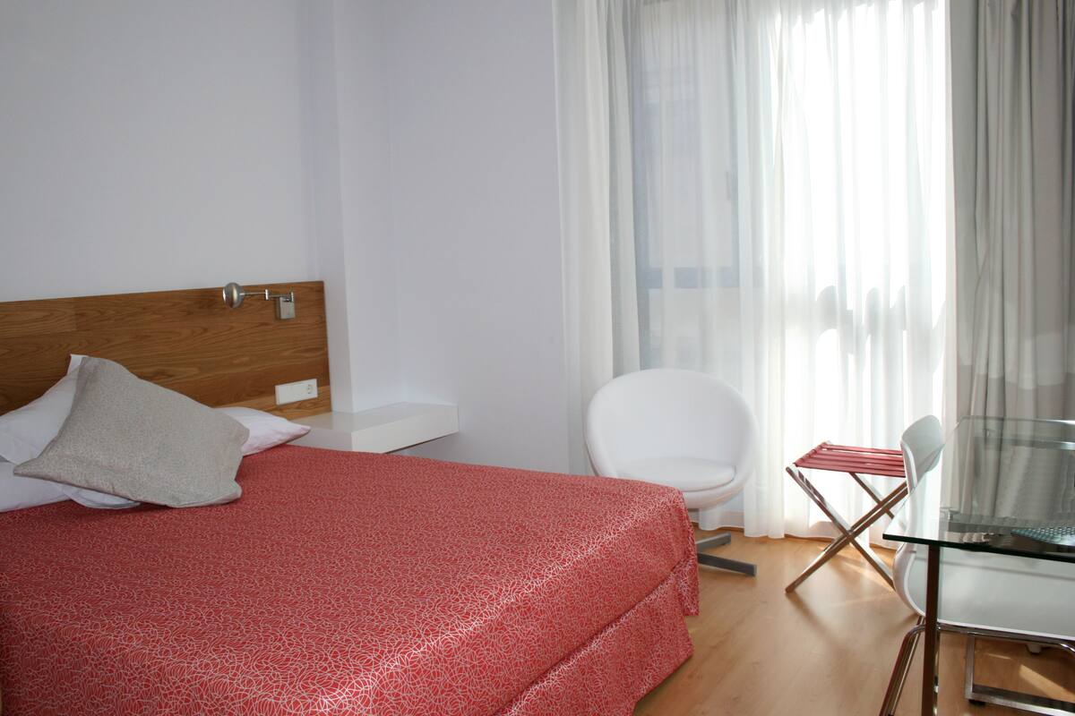 Una habitación de hotel con cama, una silla, una mesita y una ventana con cortinas.