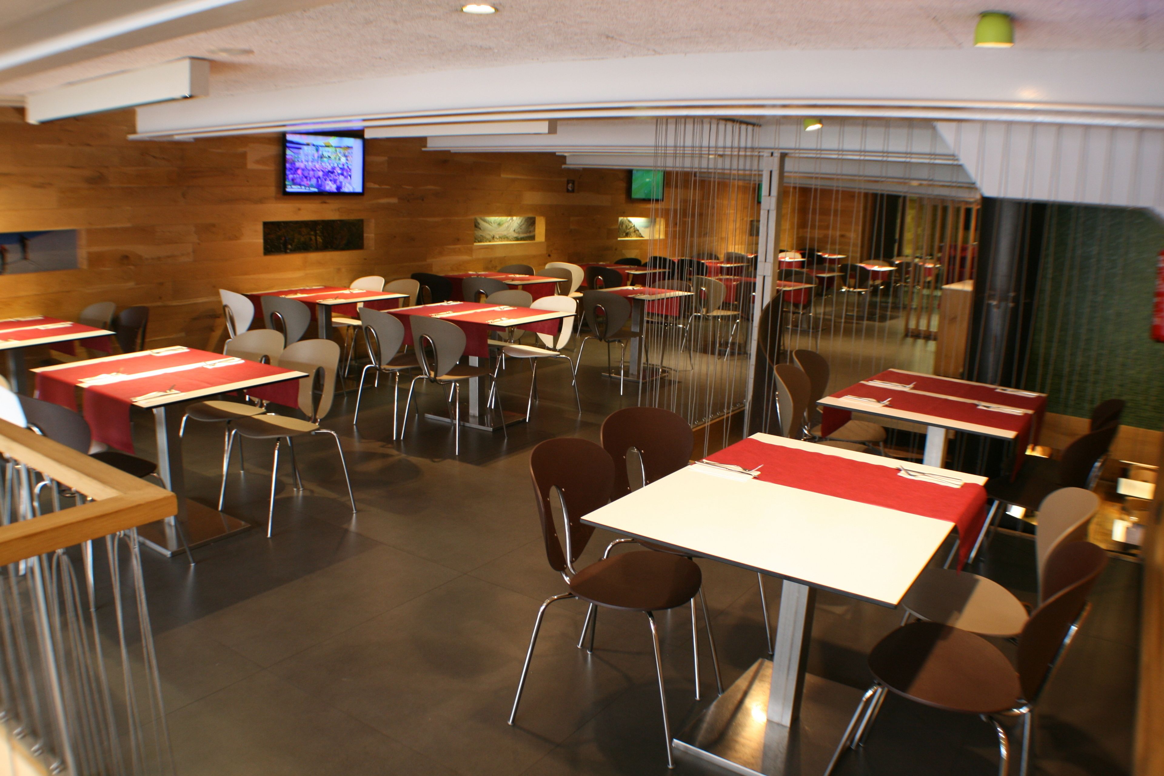 A modern restaurant with multiple tables and chairs, a flat-screen TV mounted on the wall, and a wooden accent wall.