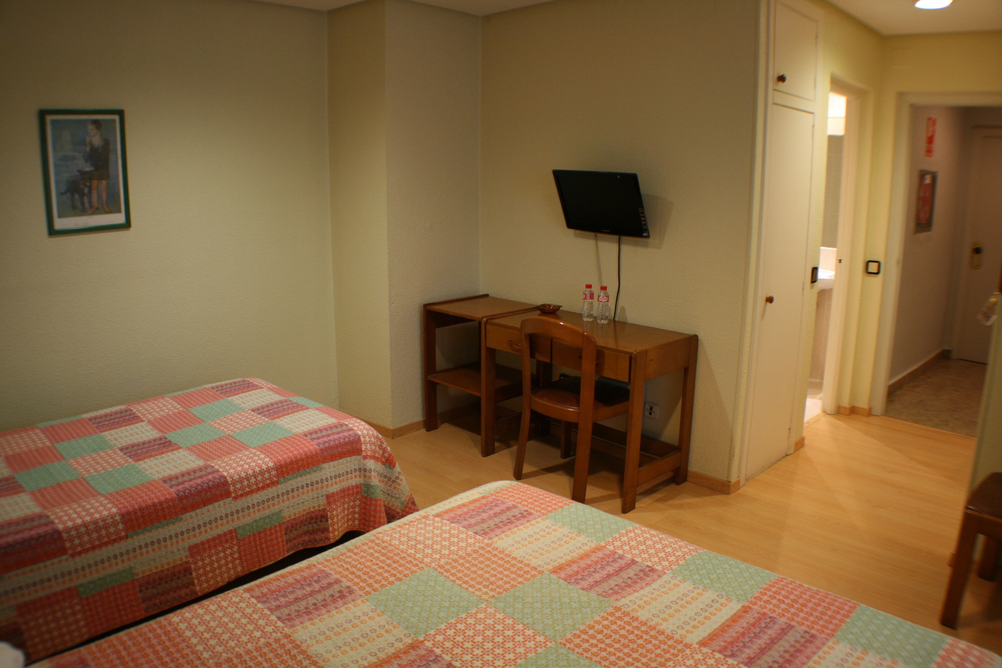 Una habitación con dos camas, un escritorio con silla, una televisión y una puerta que da a otra habitación.
