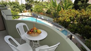 A balcony with a pool, white plastic chairs, a table with a fruit basket, and lush greenery. - Anseli Hotel Apartments and Studios (Rhodes)