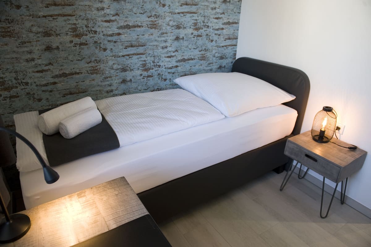 A neatly made bed with white linens and a black headboard, a bedside lamp, and a small wooden nightstand with a lamp.