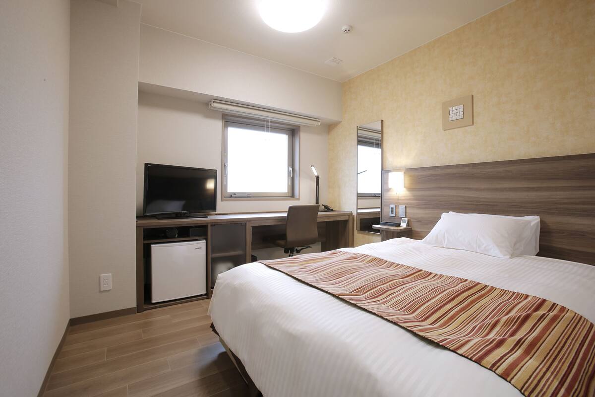 Une chambre d’hôtel comprenant un lit, un bureau avec une télévision, un petit réfrigérateur, une fenêtre avec des stores et un miroir au mur.