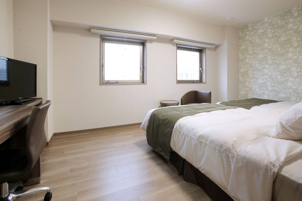 Une chambre d’hôtel avec un lit, un bureau avec un ordinateur, deux fenêtres et une chaise.