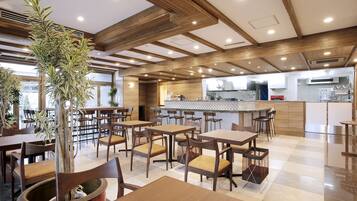 Um restaurante com mesas e cadeiras de madeira, uma cozinha ao fundo e plantas em vasos.