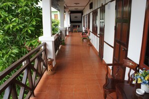 A corridor with a tiled floor, wooden railings, and a potted plant. There are chairs and a table with a vase of flowers. - Suksavanh River Hotel (Vientiane)