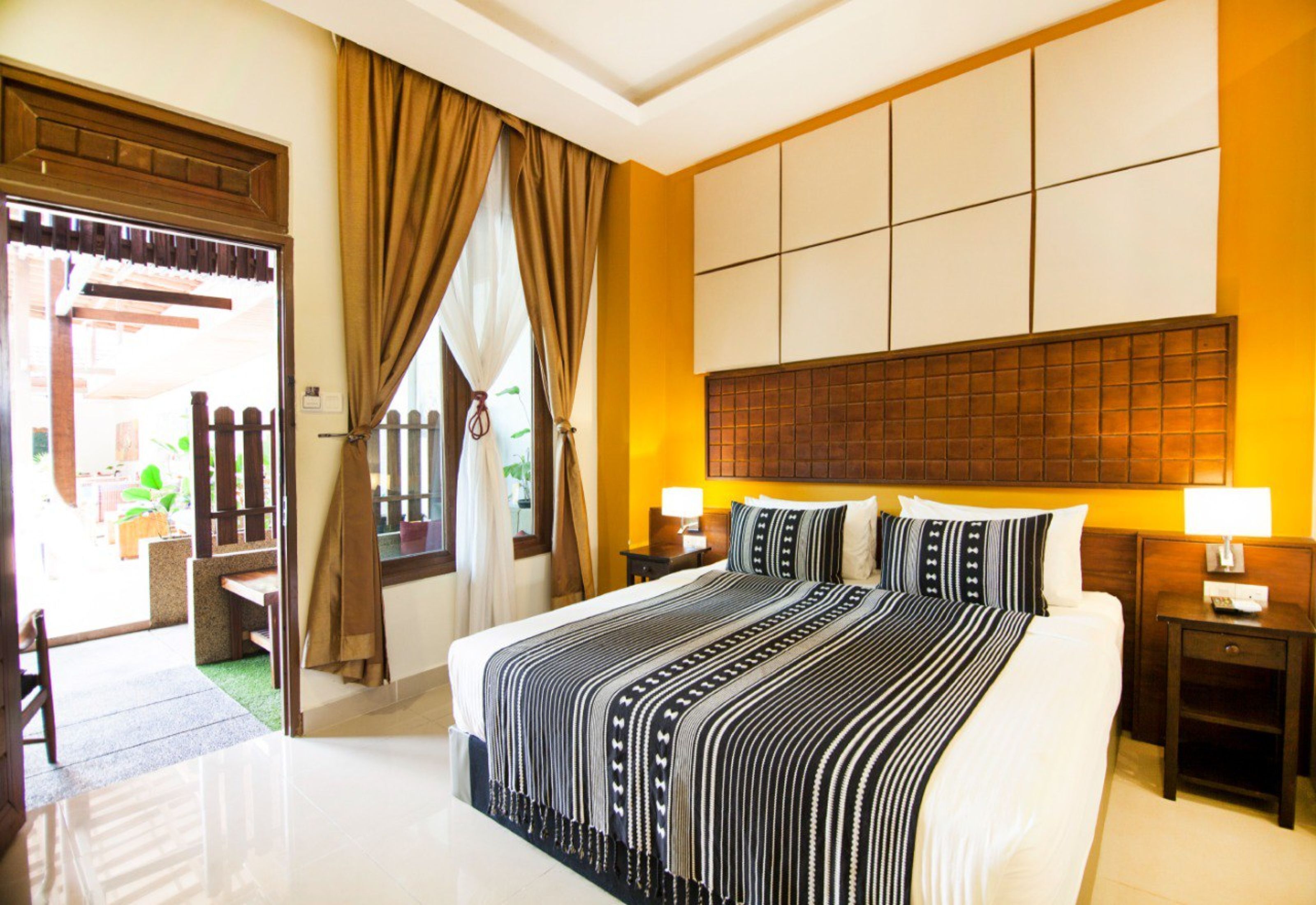 A hotel room with a large bed, two bedside tables with lamps, a wooden headboard, and a view of the outside through a door with curtains.