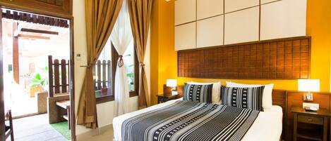 Habitación de hotel con una cama grande, dos mesitas de noche con lámparas, un cabecero de madera y vistas al exterior a través de una puerta con cortinas.