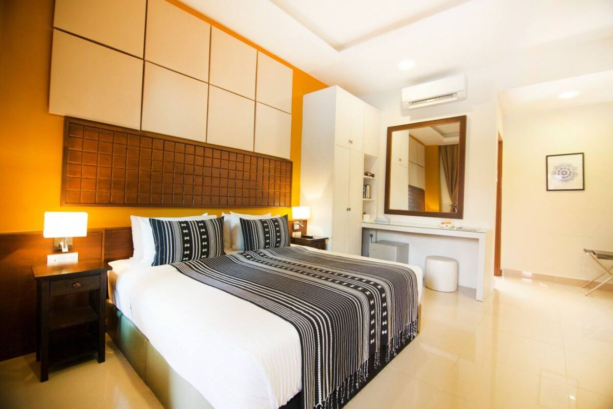 A hotel room with a large bed, bedside tables with lamps, a dresser, a mirror, and a small stool.