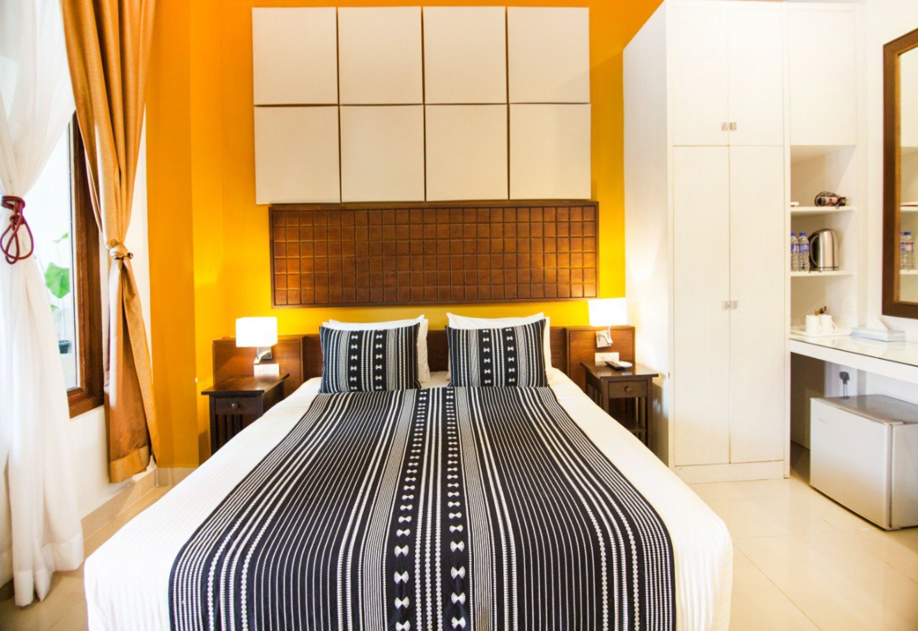 Una habitación de hotel con una cama grande, cabecera con diseño de panel, dos mesitas de noche con lámparas, un armario y una pequeña kitchenette.