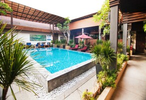 Piscina al aire libre con sillones de descanso y sombrillas, rodeada de plantas y un área techada con tejado de azulejos.