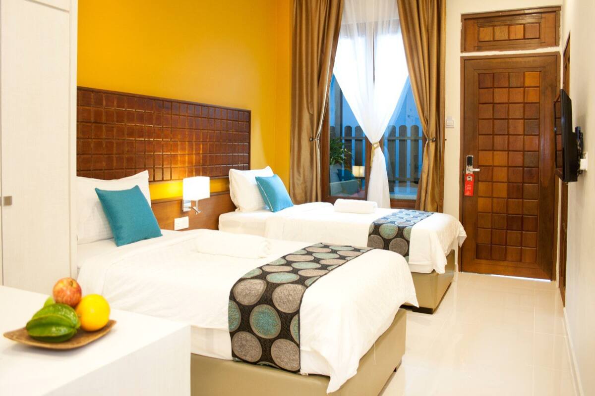 A hotel room with two beds, a wooden headboard, a bedside table with fruit, and a view of the outside.