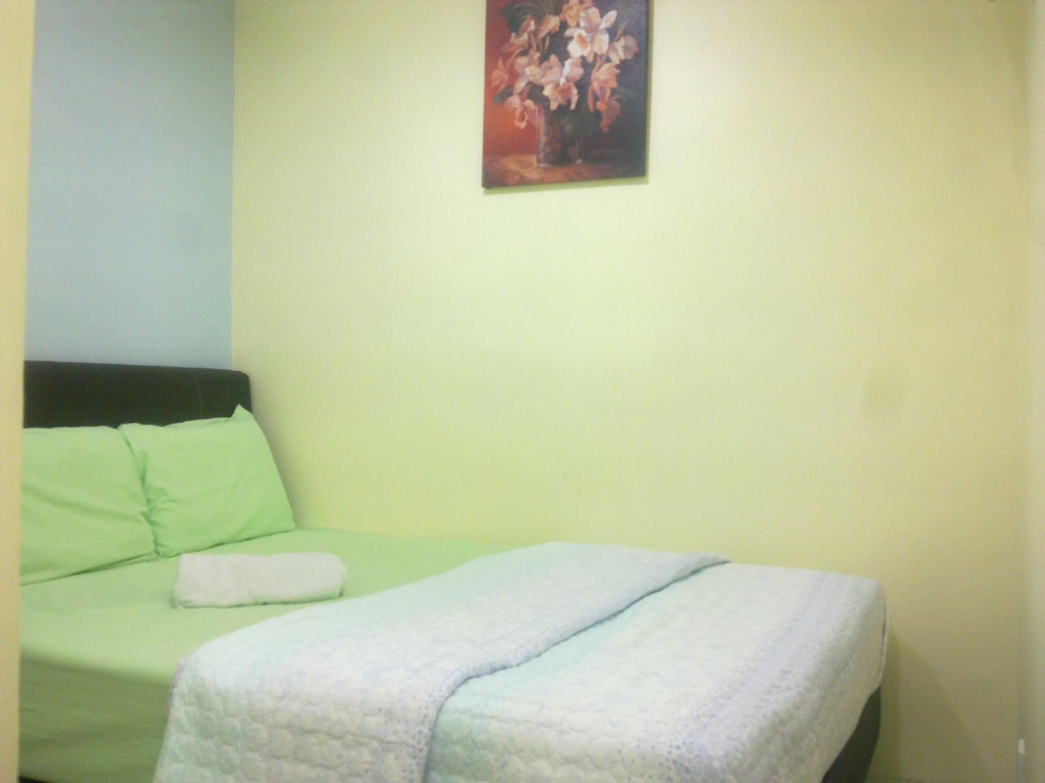 A bed with green bedding and a white blanket, a painting of flowers on the wall, and a plain light-colored wall.